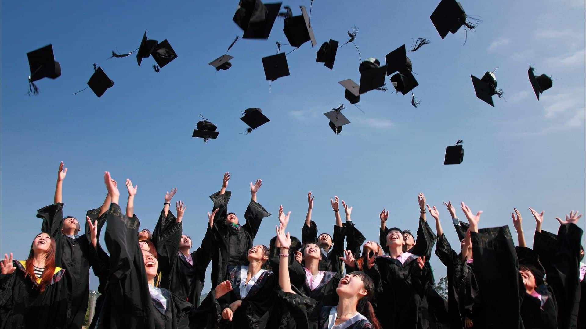 A group of university graduates throw their mortarboard hats into the air in celebration on a sunny day.
